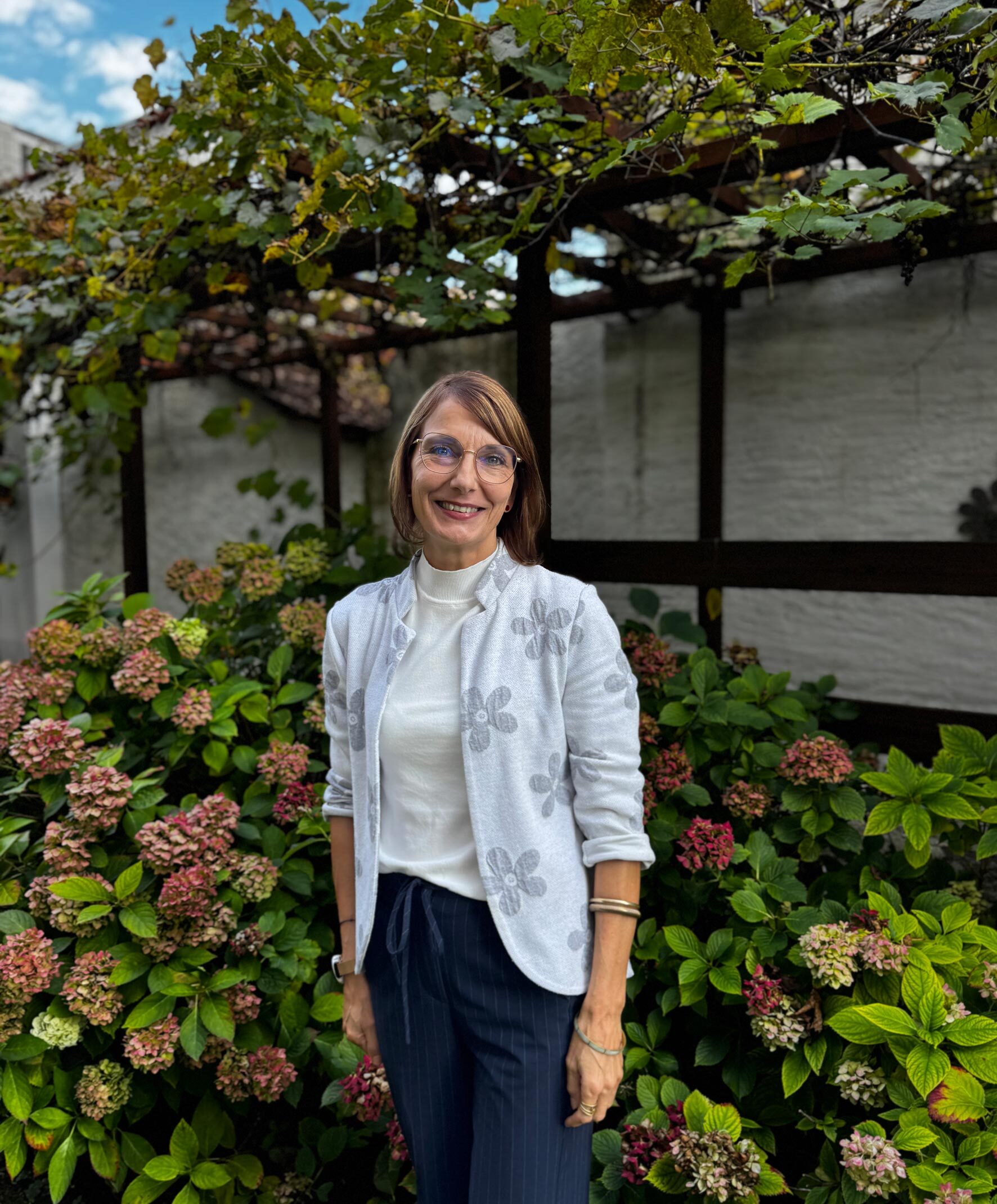 Frau mit Brille und hellem Blazer steht lächelnd im Garten vor blühenden Hortensioen und einer mit Wein bewachsenen Pergola.