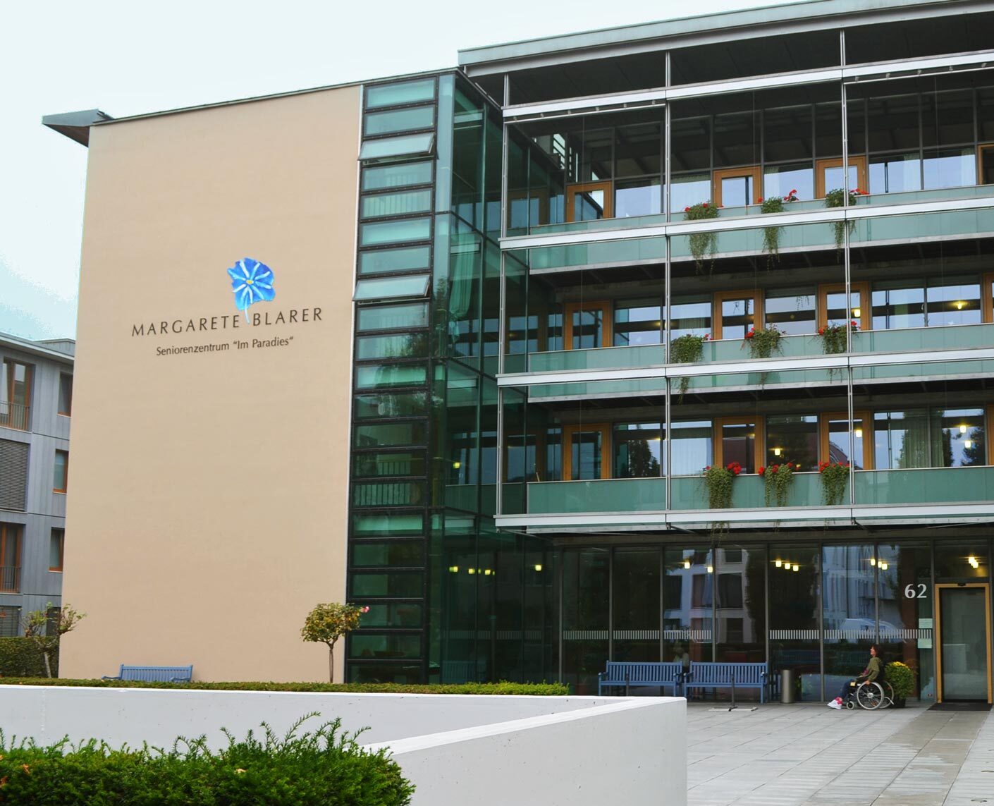 Moderne Glasfassade des Senioren­zentrums „Margarete Blarer – Im Paradies“ mit Eingangsbereich und einer Person im Rollstuhl auf dem Vorplatz.