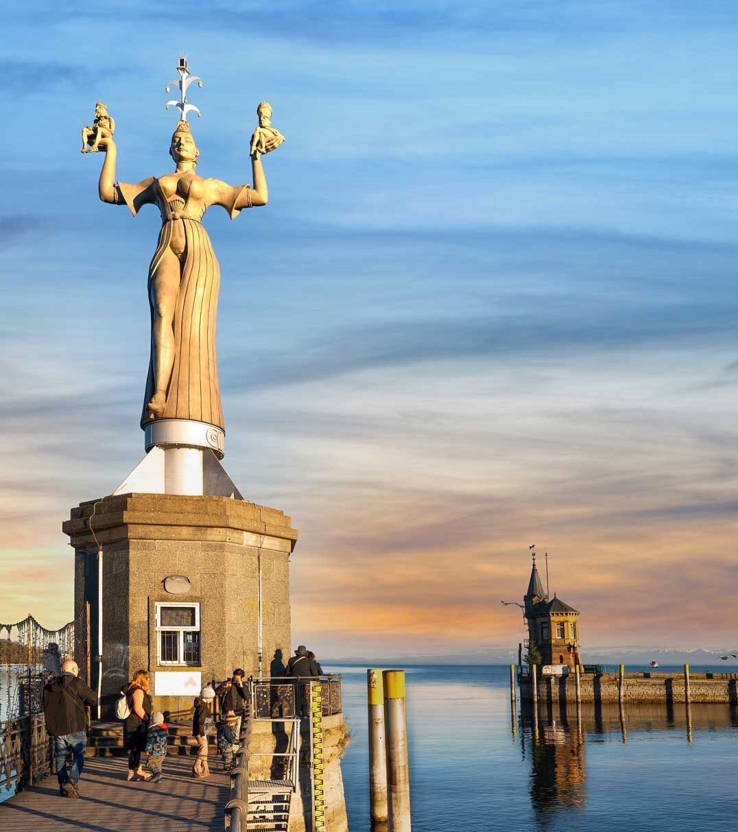 Statue "Imperia" am Hafen von Konstanz bei Sonnenuntergang, mit Menschen auf dem Holzsteg im Vordergrund und ruhigem Bodensee im Hintergrund.