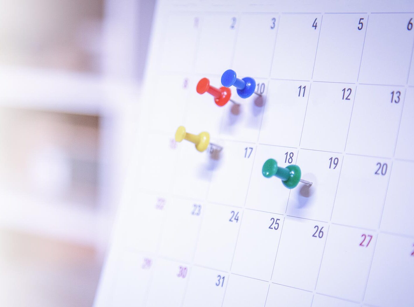 Nahaufnahme eines Wandkalenders mit mehreren bunten Pinnnadeln auf verschiedenen Tagen, symbolisch für Termineinplanungen oder Veranstaltungsübersicht.