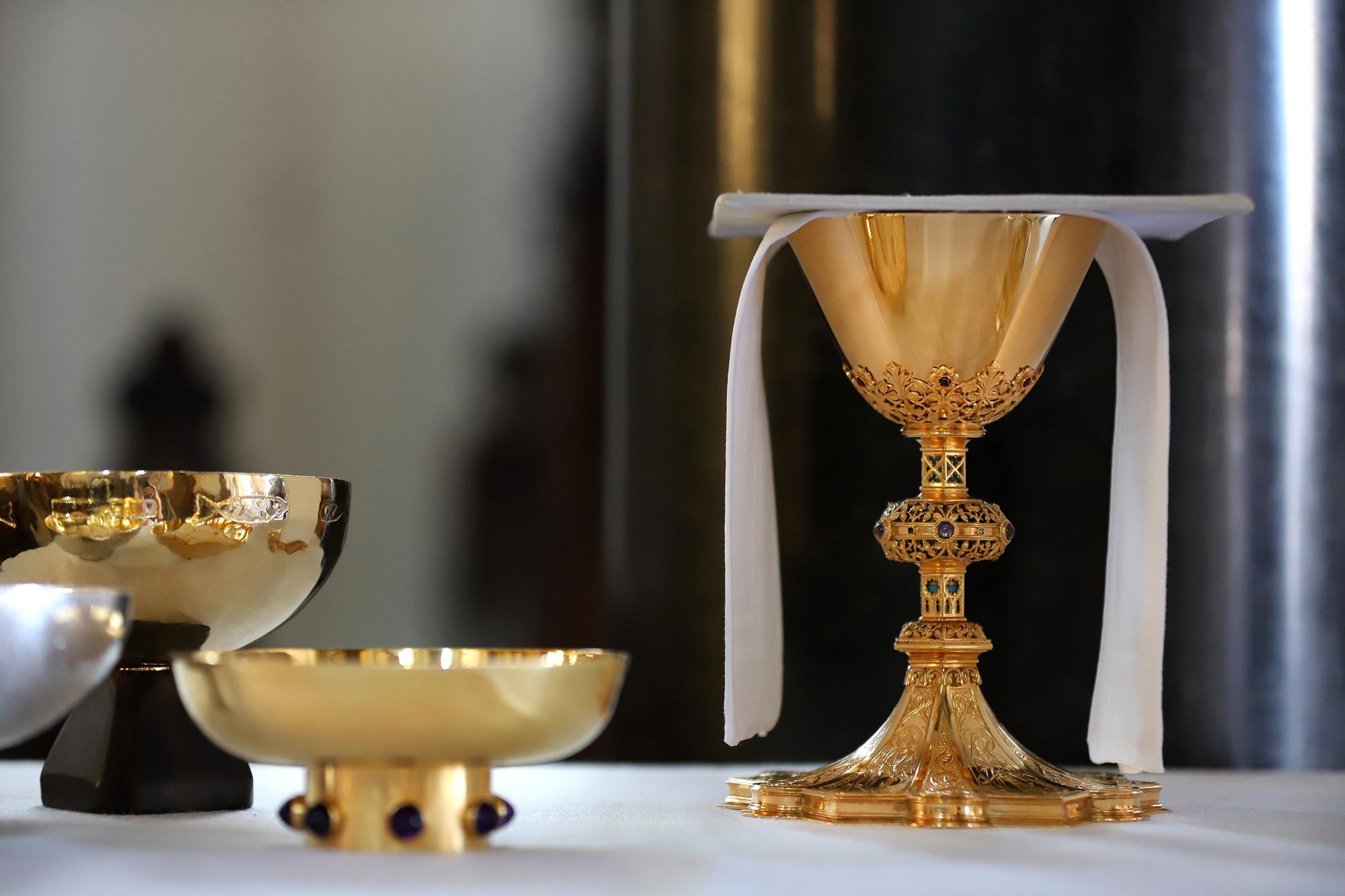 Vergoldeter, reich verzierter Kelch mit weißem Tuch, daneben weitere liturgische Gefäße auf einem Altartisch. Das Arrangement deutet auf eine katholische Messfeier oder Eucharistiefeier hin.