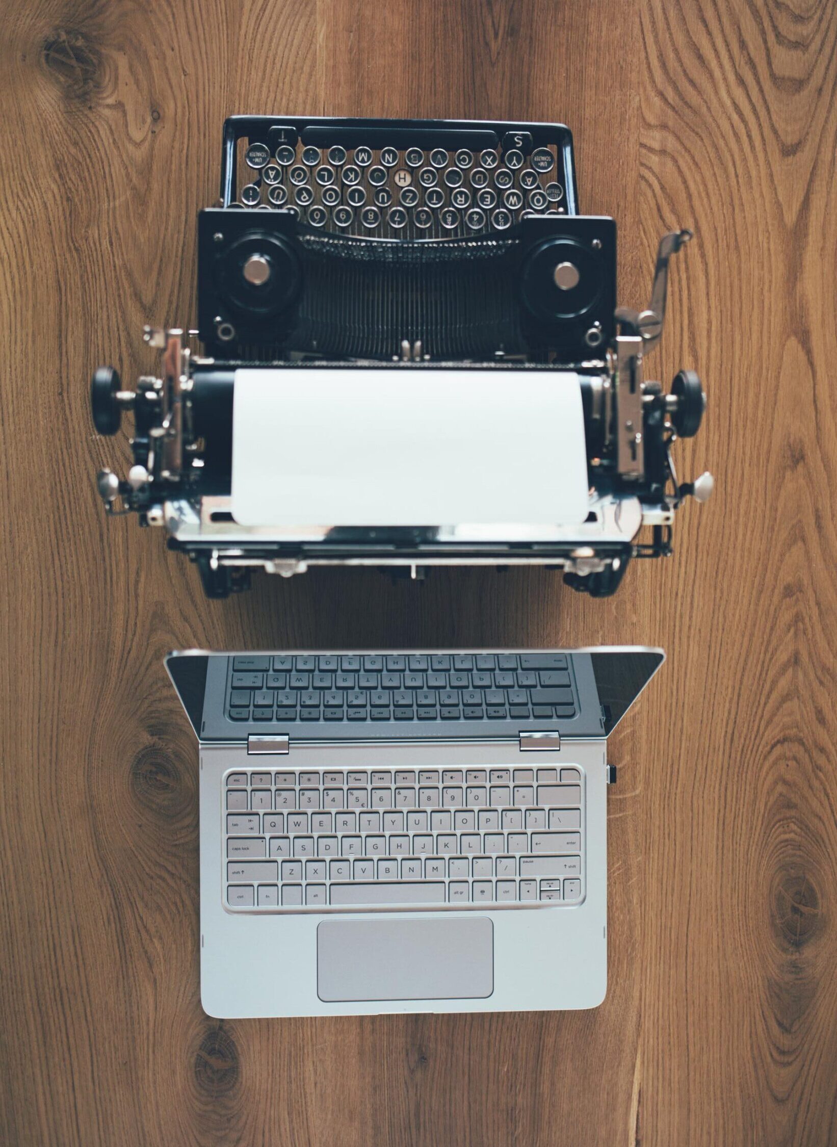 Obere Draufsicht auf einen Holztisch mit einer alten Schreibmaschine links und einem modernen Laptop rechts – symbolisch für den Wandel von analog zu digital.