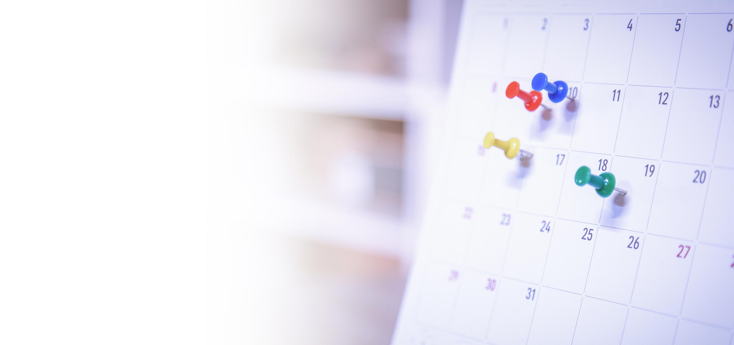 Das Bild zeigt eine Naheaufnahme eines Kalenderblatts, auf dem mehrere bunte Pinnnadeln stecken. man erkennt rote, blaue, gelbe und grüne Nadeln, die verschiedene tage markieren.