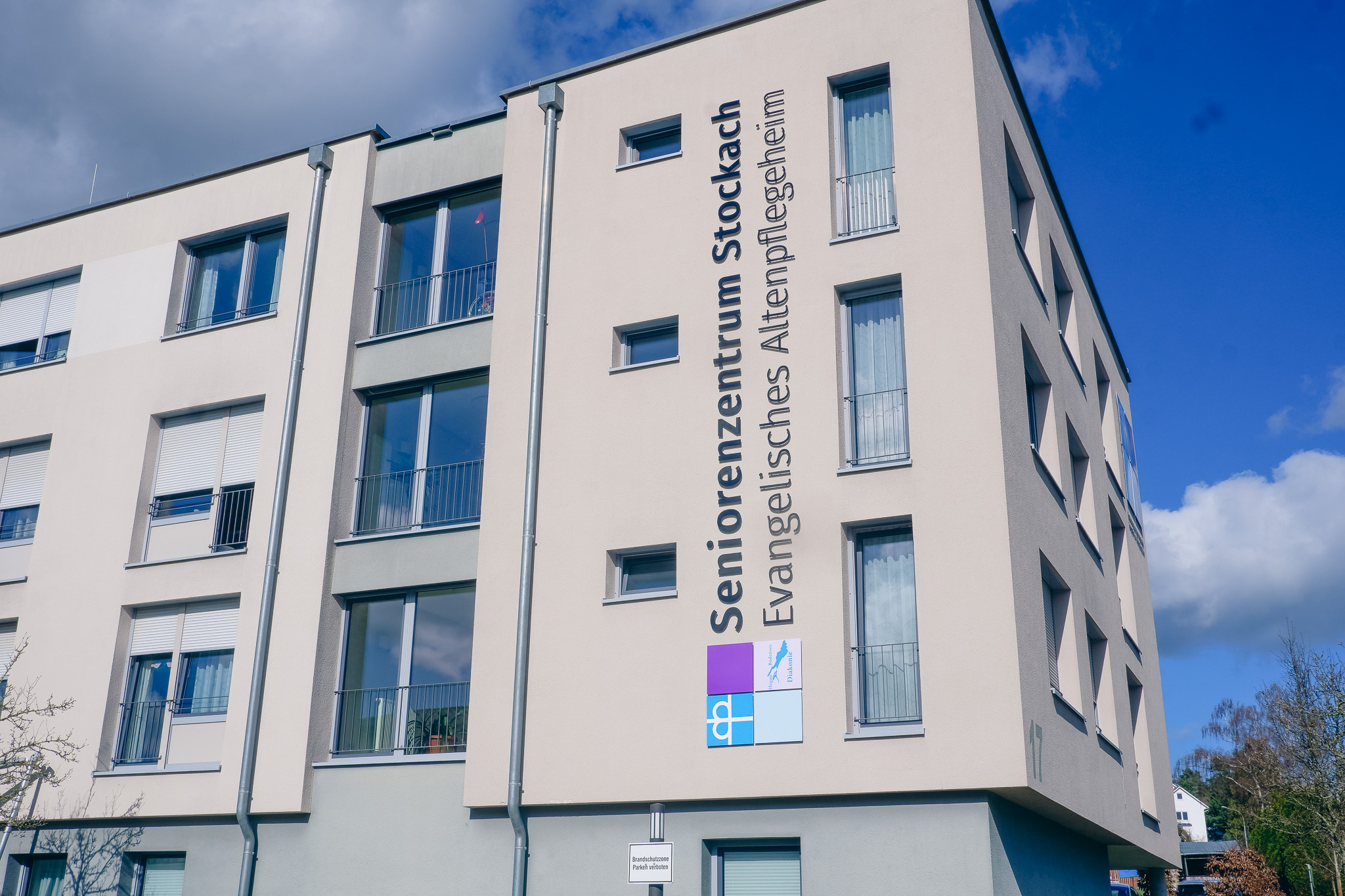 Das Bild zeigt die moderne Außenfassade des Seniorenzentrums Stockach bei hellem Tageslicht und blauem Himmel. Gebäude: Ein mehrstöckiger, heller Neubau mit einer schlichten, modernen Fassade. Beschriftung: Vertikal an der Fassade angebracht ist der Schriftzug „Seniorenzentrum Stockach – Evangelisches Altenpflegeheim“. Markenidentität: Unter dem Schriftzug sind die bekannten quadratischen Diakonie-Symbole (Kronenkreuz und Diakonie-Logo) zu sehen, was die Trägerschaft der Einrichtung verdeutlicht.