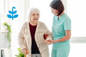 Eine Pflegerin in türkisfarbener Kleidung hilft einer älteren Frau mit weißem Haar und Gehstock beim Gehen. Beide lächeln, die Szene wirkt warm und fürsorglich.